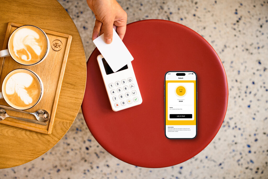 “Cafébord med två cappuccinos, en betalterminal och en iPhone som visar Föreningshäftets aktiveringssida för en deal.