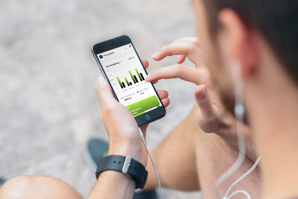 Person som sitter utomhus och håller en mobil där Föreningshäftets försäljningsstatistik visas på skärmen.