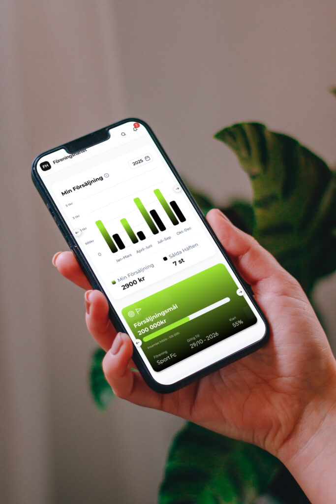 Hand som håller en mobiltelefon med Föreningshäftets personliga försäljningsstatistik på skärmen, visad framför en växt inomhus.