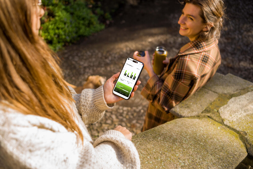 Person som håller en mobil med Föreningshäftets försäljningsstatistik på skärmen medan hon står och pratar med en vän utomhus.